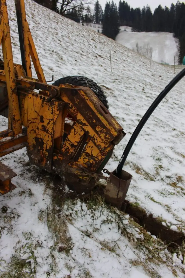 Eine Pflügmaschine arbeitet auf dem mit Schnee bedeckten Feld, während sie eine Wasserleitung im Boden verlegt. Die Pflugschare durchbrechen die winterliche Erde, um den Weg für die neue Leitung zu bahnen.